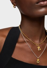 Collane a strati con una catena di perle dorate e una catena di perle argentate, ciascuna con pendenti decorativi unici che mostrano pietre colorate.