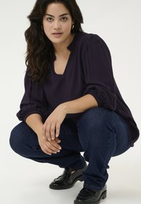 Top morado oscuro con escote redondeado y mangas gather, combinado con jeans azul oscuro y mocasines negros; en pose de cuclillas.