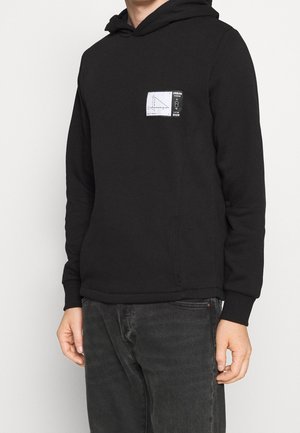 Person trägt schwarzen Hoodie mit kleinem rechteckigem Grafikdruck, der ein Diagramm und Text auf der linken Brust zeigt, kombiniert mit dunkelgrauen Jeans.