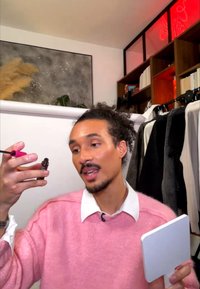 Mann in pinkem Pullover hält eine kleine Kosmetikflasche und einen Spiegel und demonstriert das Auftragen von Make-up in einem Raum mit Kleidung und Bücherregalen.