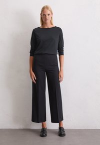 Zwarte lange mouwen top gecombineerd met hoge taille, wijde zwarte broek en zwarte instappers, geplaatst tegen een neutrale textuur achtergrond.