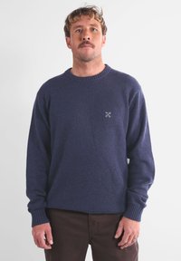 Pull en maille bleu marine avec un col rond et des poignets côtelés. Présente un petit logo blanc sur la poitrine. Matière douce et texturée.