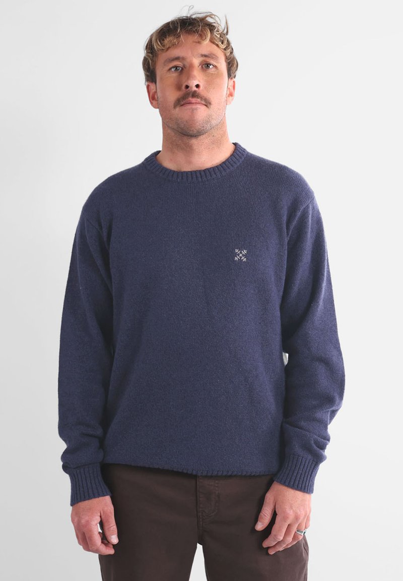 Pull en maille bleu marine avec un col rond et des poignets côtelés. Présente un petit logo blanc sur la poitrine. Matière douce et texturée.