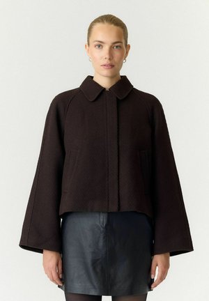 Mujer con el pelo peinado hacia atrás lleva una chaqueta de manga ancha de textura marrón oscuro y una falda de cuero negra sobre un fondo claro y liso.