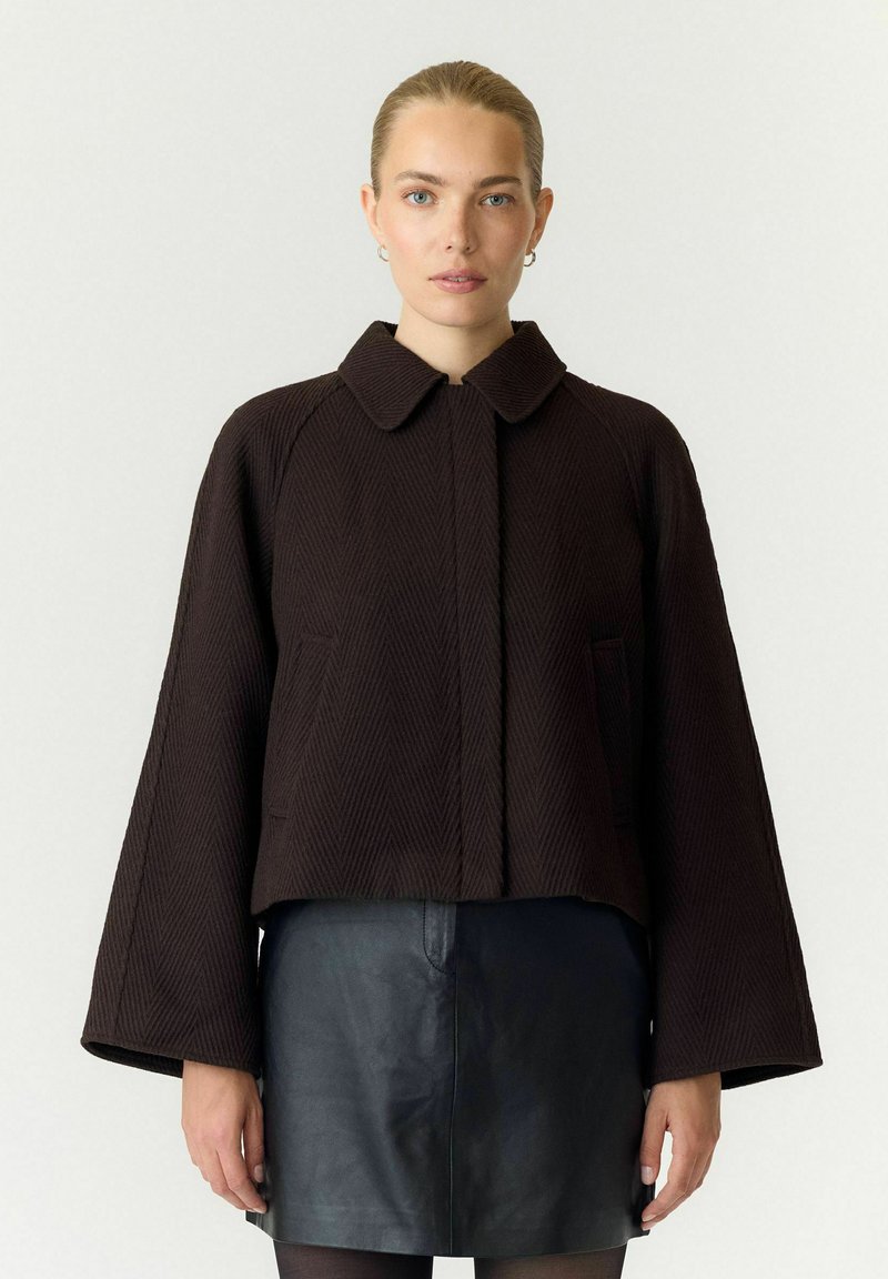 Femme aux cheveux tirés en arrière portant une veste marron foncé texturée à manches larges et une jupe en cuir noire sur un fond clair uni.