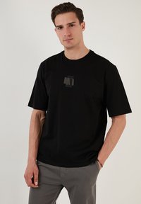 Melnas kokvilnas T-krekls ar īsām piedurknēm, kura krūtīs ir maigs pelēks "M1" logo. Standarta piegriezums ar apaļu kakla izgriezumu.
