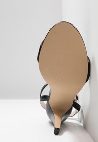 Sandale noire à talons aiguilles avec un design à bout pointu et des fines lanières. La semelle est beige avec une texture striée, présentant une finition lisse et brillante.