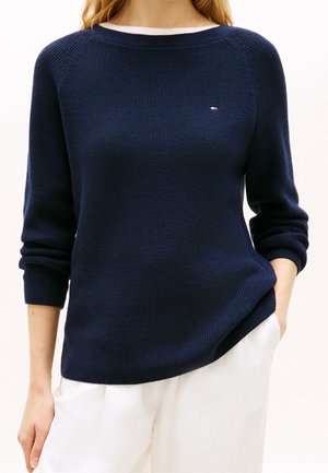 Kobieta ubrana w granatowy sweter z dzianiny z małym logo na piersi i białe spodnie, jedna ręka w kieszeni, na jednolitym tle.