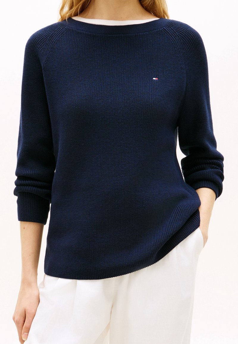 Femme portant un pull en tricot bleu marine avec un petit logo sur la poitrine et un pantalon blanc, une main dans la poche, sur un fond uni.
