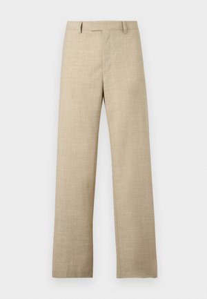 Młody mężczyzna ubrany w beżowe krótki rękaw dzianinowa polo, szerokie beżowe spodnie, czarne skórzane buty i czarną torbę na ramię, stoi na białym tle.