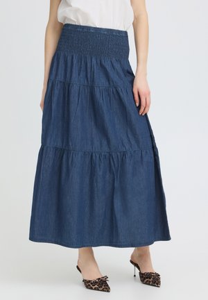 Naine kannab pikkade astmeliste tumel sinisel teksasekil koos valge topi ja leopardimustriliste teravate kontsadega kingadega, seisab ühevärvilise tausta ees.