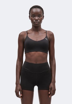 Slank kvinde iført sort Puma sports-bh og højtaljede sorte træningsshorts, stående mod en almindelig lys baggrund.