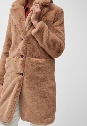 Personne portant un long manteau marron en fausse fourrure douce avec des boutons, tenant le col d'une main, sur un fond clair uni.