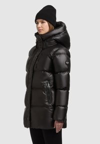 Fekete, fényes, sima anyagú pufi dzseki kapucnival és cipzáras zsebekkel. Testhezálló szabás és feltűnő varrási részletek jellemzik.