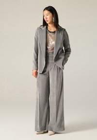 Completo in velluto a coste grigio chiaro con pantaloni a gamba larga e blazer a un bottone, indossato sopra un top con paillettes. Presenta sottili righe verticali e orecchini eleganti.