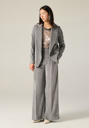 Completo in velluto a coste grigio chiaro con pantaloni a gamba larga e blazer a un bottone, indossato sopra un top con paillettes. Presenta sottili righe verticali e orecchini eleganti.