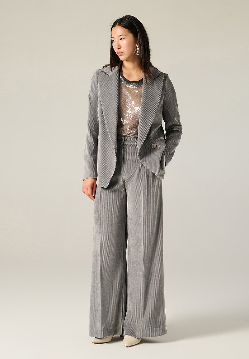 Completo in velluto a coste grigio chiaro con pantaloni a gamba larga e blazer a un bottone, indossato sopra un top con paillettes. Presenta sottili righe verticali e orecchini eleganti.