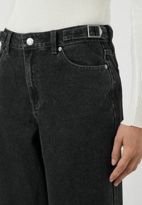 Los jeans de mezclilla negra presentan un diseño de cintura alta, cinco bolsillos y una textura elegante con un suave efecto de lavado. Cierre de botón metálico como acento.