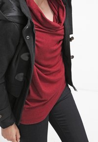 Haut rouge à col montant en tissu doux, associé à un manteau duffle noir avec des brandebourgs et une texture lisse. Pantalon noir ajusté.