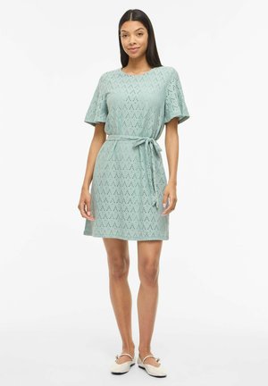 Jeune femme debout portant une robe bleu clair à manches courtes avec une ceinture à la taille et des chaussures plates blanches sur fond blanc.