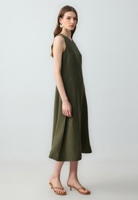 Robe sans manches vert olive avec une silhouette fluide, présentant des coutures en dart et une coupe au genou. Associée à des sandales à talons beiges.