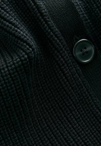 Zwart gebreid sweaterstof met een geribbelde textuur en een zichtbare zwarte viergaatjesknop. Het ontwerp richt zich op een close-up van de materiaaldetails.