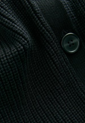 Zwart gebreid sweaterstof met een geribbelde textuur en een zichtbare zwarte viergaatjesknop. Het ontwerp richt zich op een close-up van de materiaaldetails.