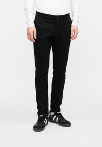 Muž v priliehavých čiernych nohaviciach, bielej košeli s dlhým rukávom a čiernych teniskách s bielymi pruhmi na bielom pozadí.