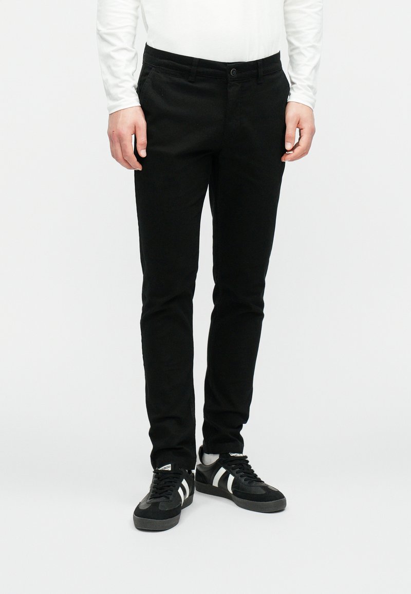 Muž v priliehavých čiernych nohaviciach, bielej košeli s dlhým rukávom a čiernych teniskách s bielymi pruhmi na bielom pozadí.