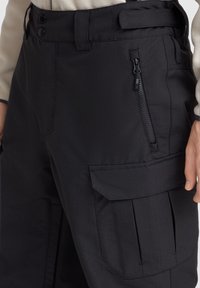 Musta cargo-housut, joissa on sileä pinta, vetoketjullinen tasku, napitus ja sivutasku läpällä.