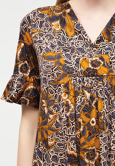 Γυναίκα που φοράει φόρεμα με V-λαιμόκοψη και κοντά μανίκια με βολάν, με λεπτομερή λουλουδάτο και αφηρημένο μοτίβο σε μουσταρδί, ναυτικό μπλε και λευκά χρώματα.