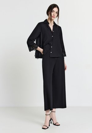 Blusa nera con bordi in pizzo sulle maniche a campana, abbinata a pantaloni neri a gamba larga e sandali neri con tacco alto e cinturini. Tessuto dal tocco morbido.