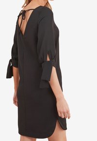 Femme portant une robe noire, longueur genou, avec des manches trois-quarts nouées au niveau des coudes et un dos nu attaché par un nœud au cou.