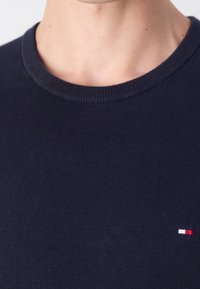 Gros plan sur une personne portant un pull en laine bleu marine avec un petit logo rectangulaire rouge, blanc et bleu sur la poitrine.