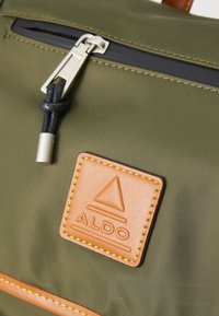 Sac à dos en nylon vert olive avec un patch en cuir tan. Il dispose d'une fermeture éclair argentée avec un cordon noir et des coutures jaunes autour du patch.