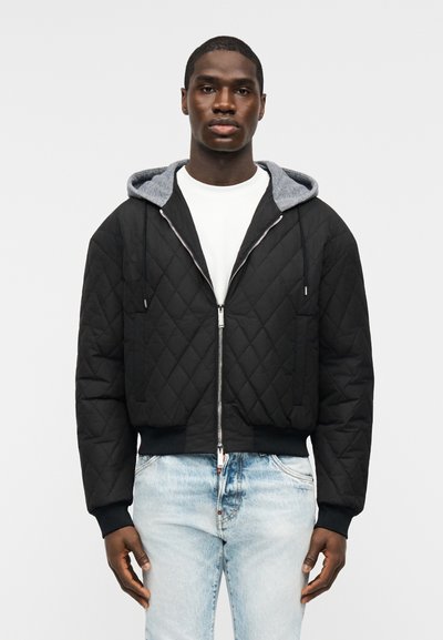 Homme portant une veste noire matelassée zippée avec une capuche grise, une chemise blanche et un jean bleu clair, debout devant un fond uni.