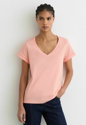 Joven con el cabello trenzado que lleva una camiseta de manga corta color rosa claro con cuello en V y jeans oscuros, de pie contra una pared blanca lisa.