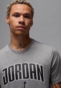 T-shirt grigia con scollo rotondo caratterizzata da un grande logo "JORDAN" in nero con bordi bianchi. Tessuto morbido, vestibilità casual, indossata con collane sovrapposte.