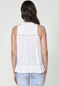 Top bianco senza maniche con tessuto testurizzato, dettagli in pizzo sulla schiena e orlo a volants. Indossato con jeans azzurri chiari.