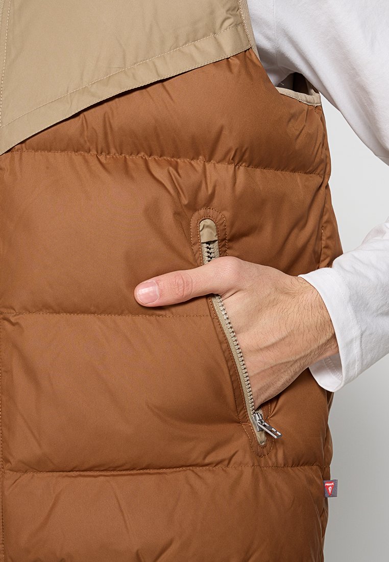 Gilet rembourré marron avec une partie supérieure beige, poche latérale zippée et texture lisse, présentant des coutures visibles et une étiquette de marque.