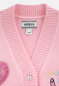 Cardigan en tricot rose clair avec un col en V, des bords côtelés, un patch en forme de cœur, un texte brodé et des détails de bouton en forme de cœur. Étiquette de marque visible.