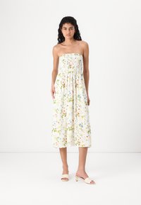 Φλοράλ φόρεμα χωρίς τιράντες με σμιλευμένο μπούστο σε λευκό χρώμα, που διαθέτει πράσινα φύλλα και πολύχρωμα λουλούδια; μεσαίου μήκους κόψιμο; το μοντέλο φορά μπεζ μπαλαρίνες.