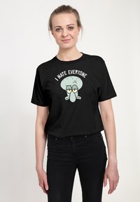 Sort t-shirt lavet af bomuld med en tegneseriefigur med et lysblåt hoved og briller, teksten lyder: "JEG HADER ALLE" ovenfor.