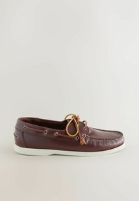 Chaussure bateau en cuir marron pour homme avec lacet jaune noué en nœud et semelle en caoutchouc blanche, vue de profil sur un fond uni.