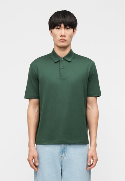 Jeune homme portant un polo à manches courtes vert foncé et un jean bleu clair, debout devant un fond blanc uni.
