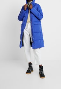 Blue puffer coat with a hood, zip front, and patch pockets. Paired with white cropped pants and black lace-up boots with rubber soles.