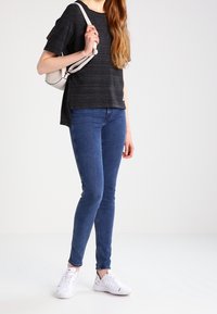 Black short-sleeve top, paired with slim-fit blue jeans and white sneakers. The model carries a small white handbag, against a neutral background.