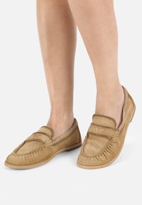 Beige Wildleder-Mokassins mit runder Zehenpartie, Quasten-Detail um die Kanten und einer glatten, flachen Sohle.