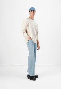 Sweatshirt beige surdimensionné, jeans bleu clair et chaussures noires ; modèle portant une casquette bleue ; tenue décontractée, design simple, coupe ample.
