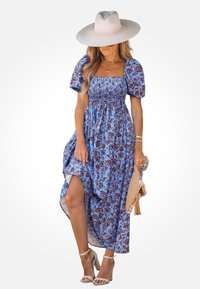 Abito maxi blu a fiori con maniche a palloncino, corpetto arricciato e spacco alto fino alla coscia. Accentuato da un cappello a tesa larga e sandali con cinturini.